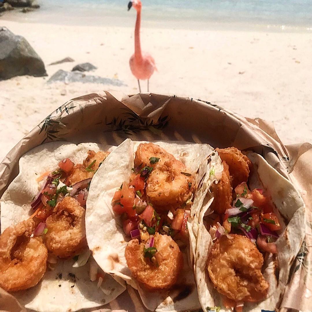 3 fried shrimp tacos with flamingo in the background on the beach. 